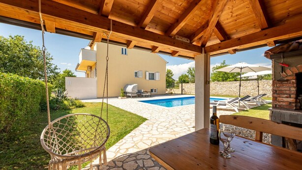Una pérgola de madera con una hamaca colgante, un patio de piedra, una piscina y un horno de ladrillo.