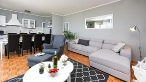 Un salon moderne avec un canapé gris, un coin repas avec des chaises noires, une table basse blanche agrémentée de fleurs et d’une bouteille, et une cuisine en arrière-plan.