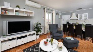 Un salon moderne doté d’un téléviseur à écran plat, d’un fauteuil gris, d’un coin repas avec une table et des chaises, et d’une cuisine avec des meubles blancs.