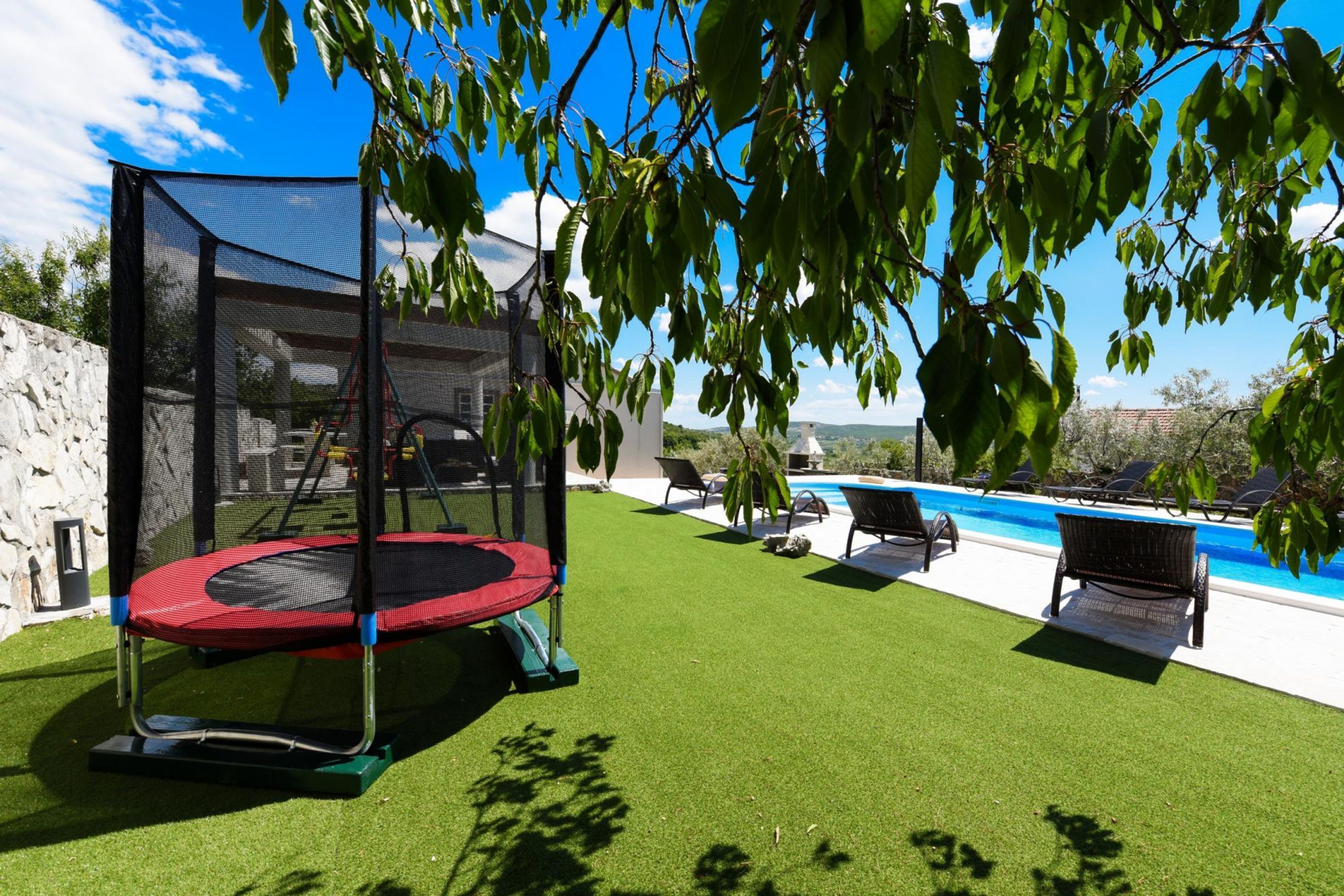 Un patio trasero con un trampolín, césped artificial, piscina y sillas de descanso.