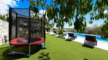 Un patio trasero con un trampolín, césped artificial, piscina y sillas de descanso.