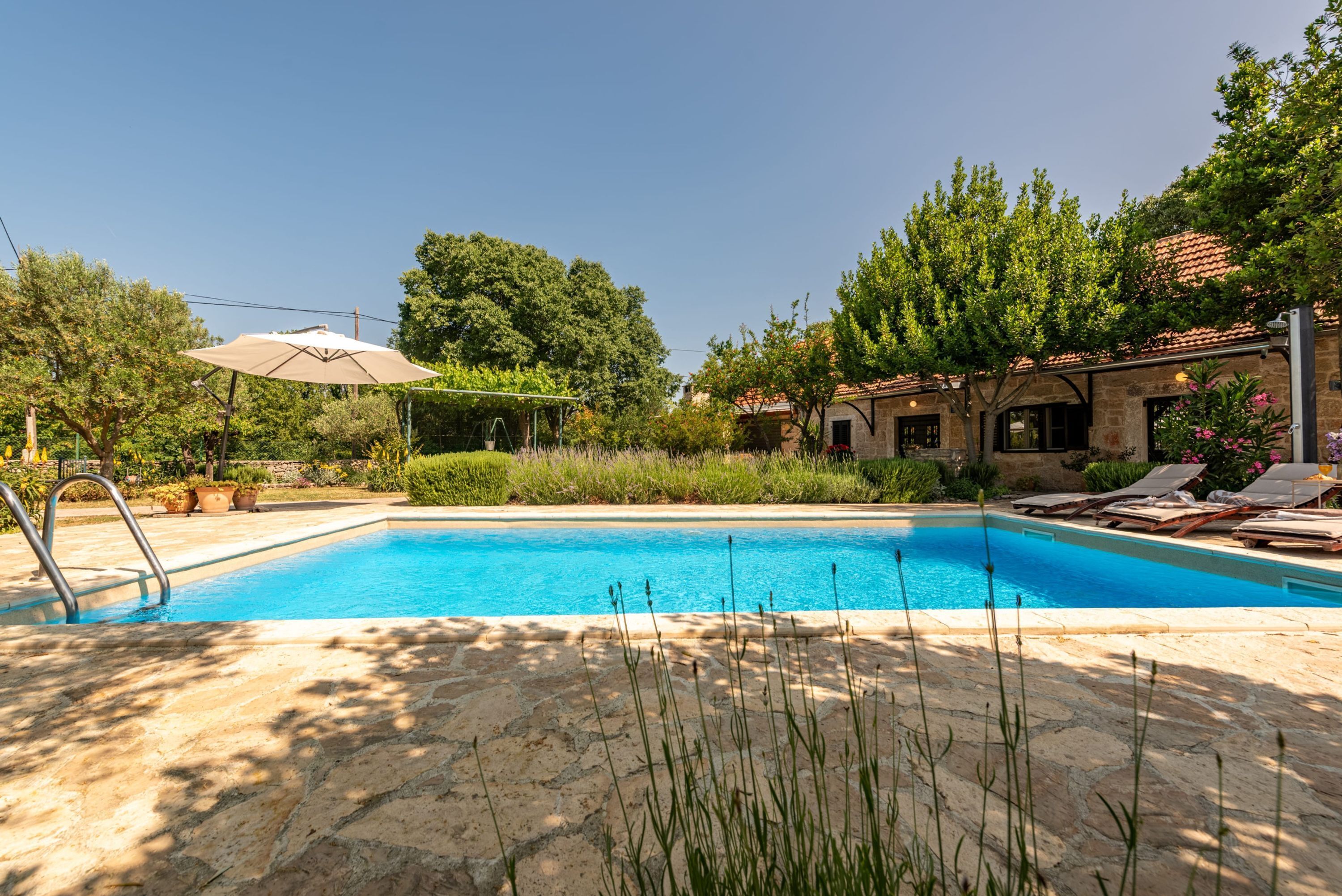 Una piscina con solárium de piedra, rodeada de vegetación y un edificio con tejado de azulejos.