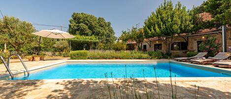 Una piscina con solárium de piedra, rodeada de vegetación y un edificio con tejado de azulejos.