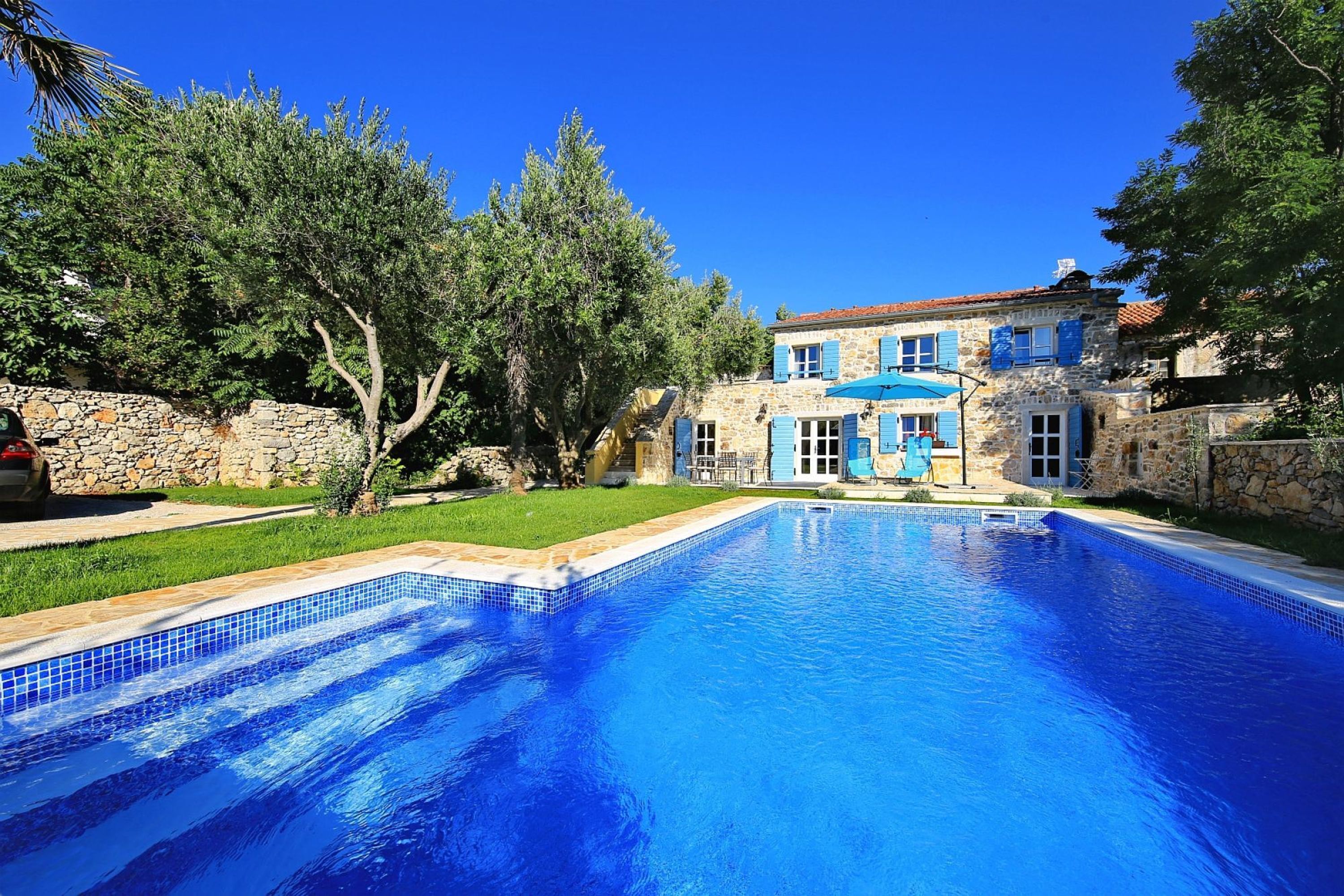 A swimming pool with a stone wall and a stone building with blue shutters in the background.