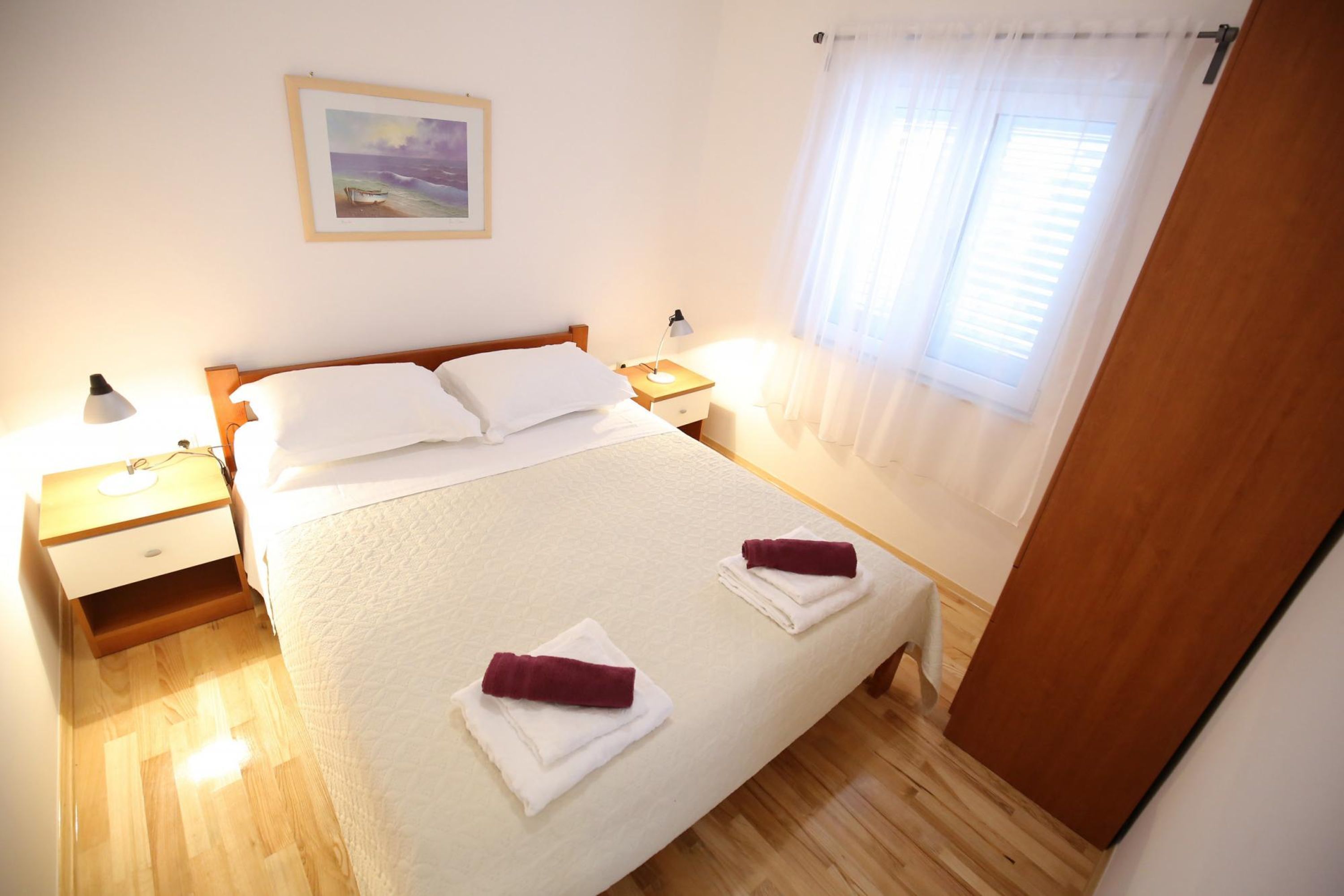 Una cama bien hecha con sábanas blancas y dos toallas dobladas, una mesita de noche con una lámpara, un cuadro en la pared y una ventana con cortinas de gasa.