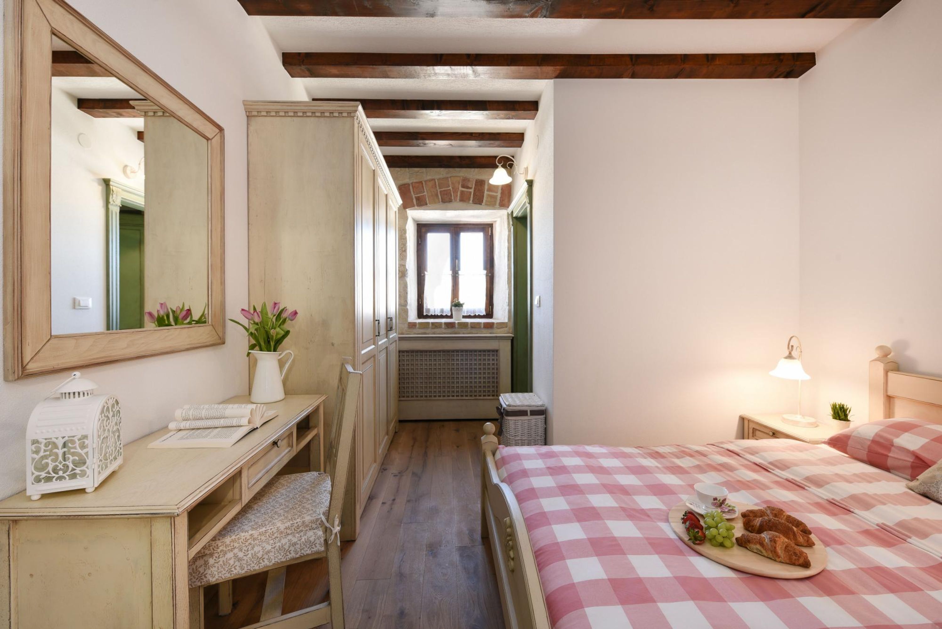 A bedroom with a checkered bedspread, a desk with a lamp, a mirror, and a window with a view of the street.