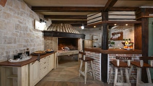 A rustic kitchen with a stone wall, wooden beams, and a brick oven. There are bar stools and a counter with a sign that reads "PRIKA". - Villa Laurana Zadarvillas (Pakostane)