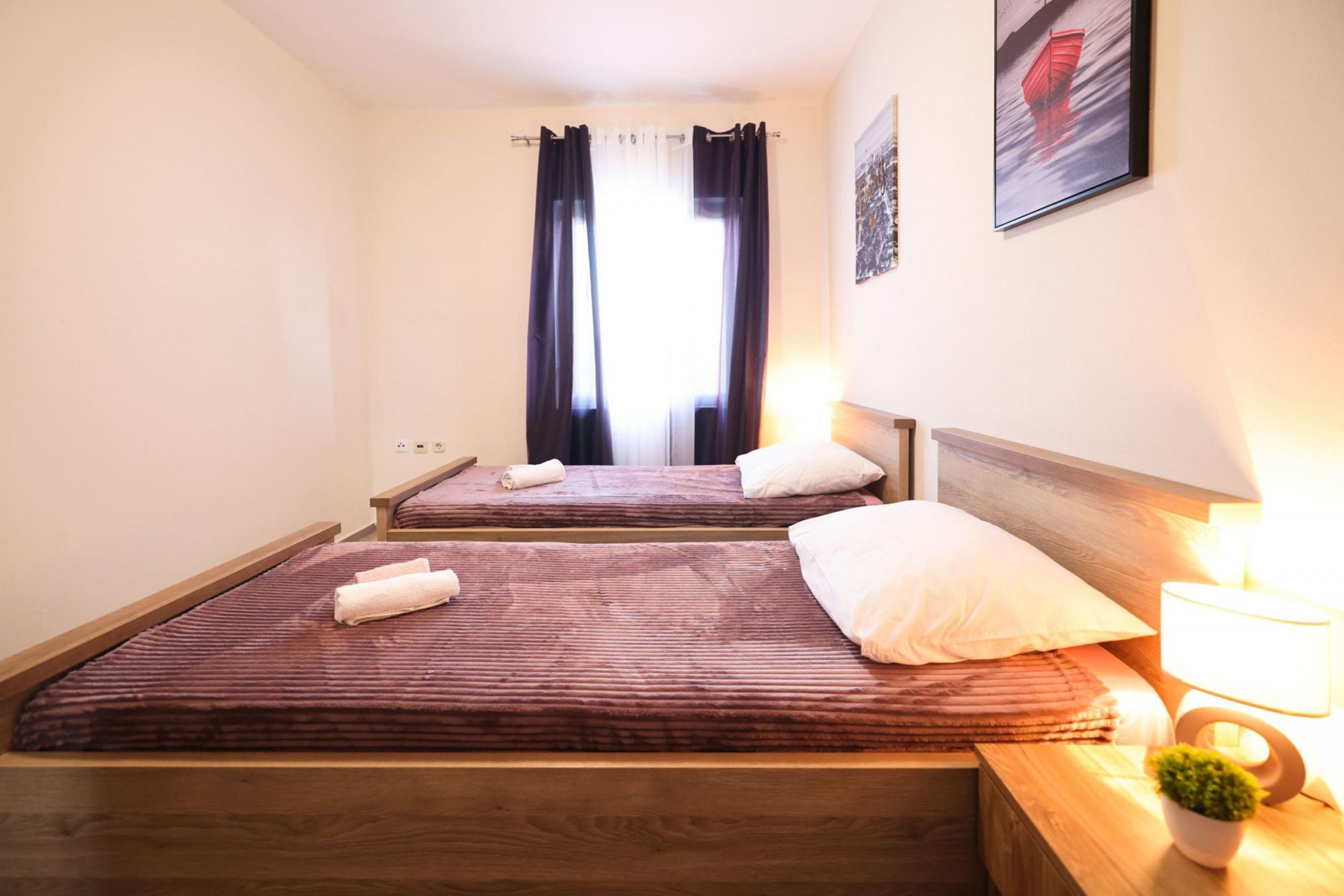 A hotel room with two beds, a wooden headboard, a nightstand with a lamp, a small potted plant, and a window with curtains.