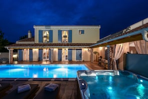 Une piscine avec un jacuzzi, un patio couvert et une maison avec un toit en tuiles et des volets.