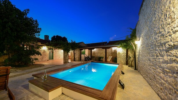 Une piscine rectangulaire avec une terrasse en bois, entourée de murs en pierre et dotée d’un espace de détente abrité.
