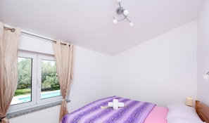 A bedroom with a bed, a window with curtains, a bedside lamp, and a view of a swimming pool.