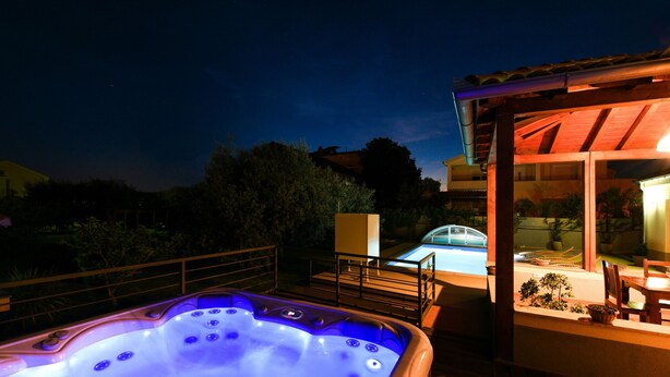 Ein Whirlpool mit Glaskonstruktion, ein Schwimmbecken und eine überdachte Terrasse mit Sitzgelegenheiten.
