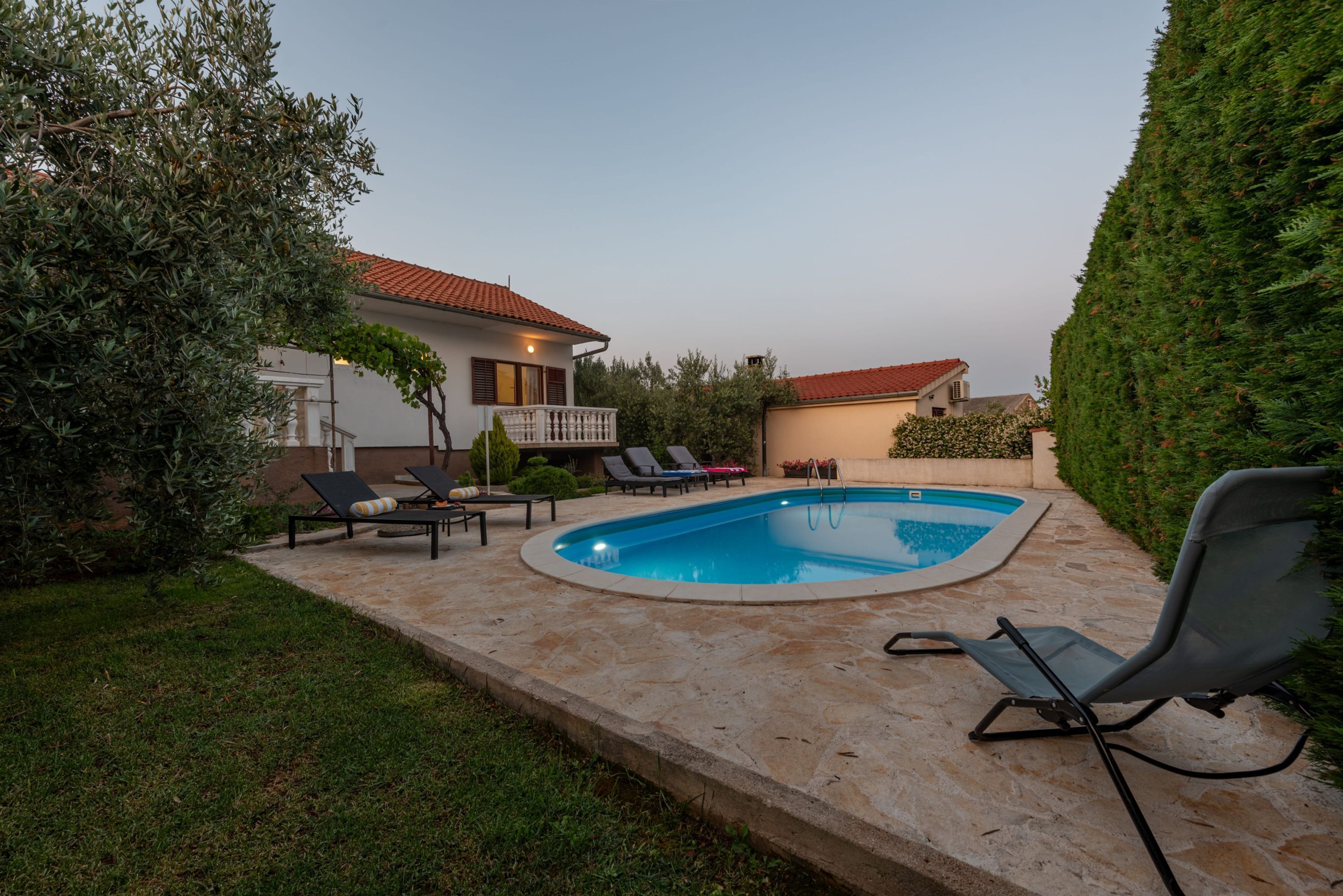 Une piscine avec des chaises longues, une terrasse en pierre et une maison avec un toit rouge en arrière-plan.
