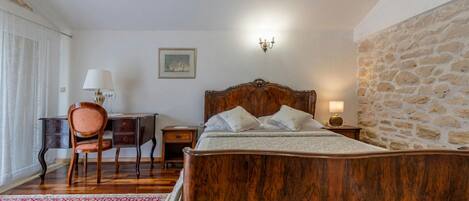 Un dormitorio con una cama de madera, un escritorio, una silla, una lámpara, un cuadro en la pared y una pared de piedra.