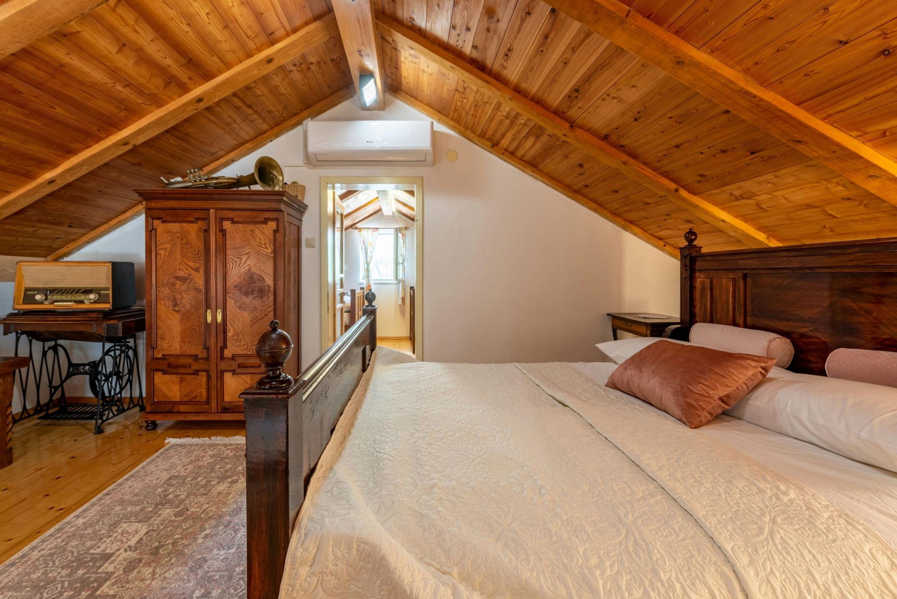 A bedroom with a sloped ceiling, wooden beams, a bed with white bedding, a wooden wardrobe, and a sewing machine.
