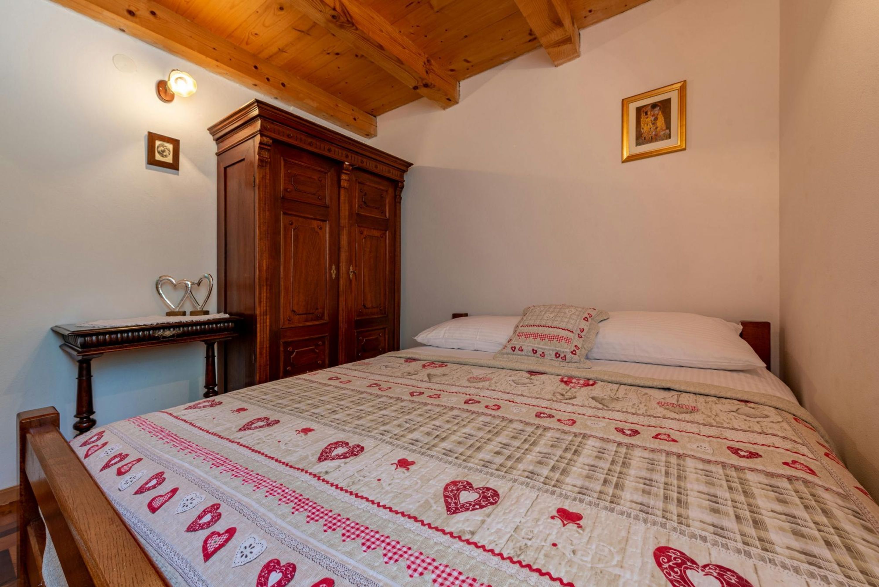 A bedroom with a wooden ceiling, a bed with a patterned quilt, a wooden wardrobe, and a small table with heart-shaped decorations.