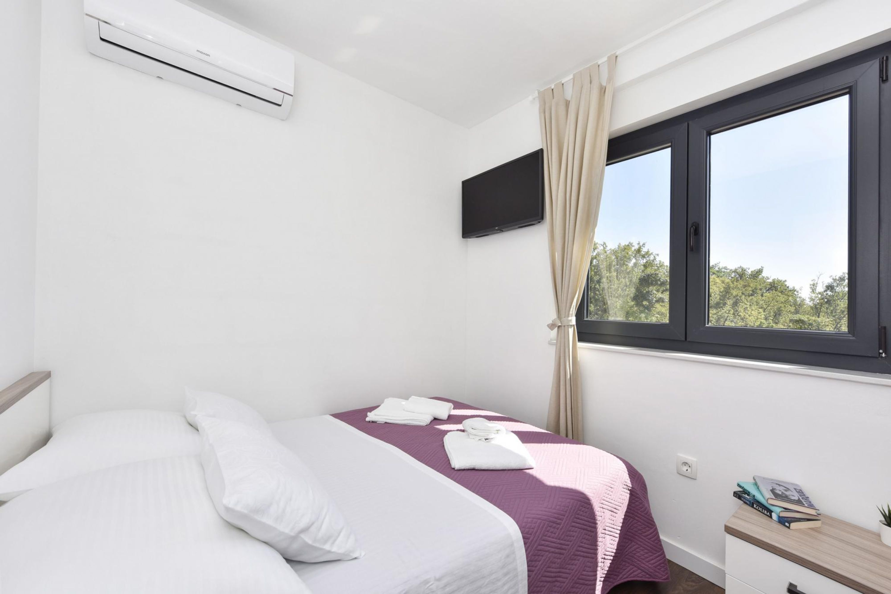 A bedroom with a bed, a TV mounted on the wall, a window with curtains, and a bedside table with books.