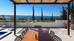 A covered outdoor dining area with a view of the sea, a pool, and a wooden table with chairs. - Villa Royal Promons Zadarvillas (Starigrad)