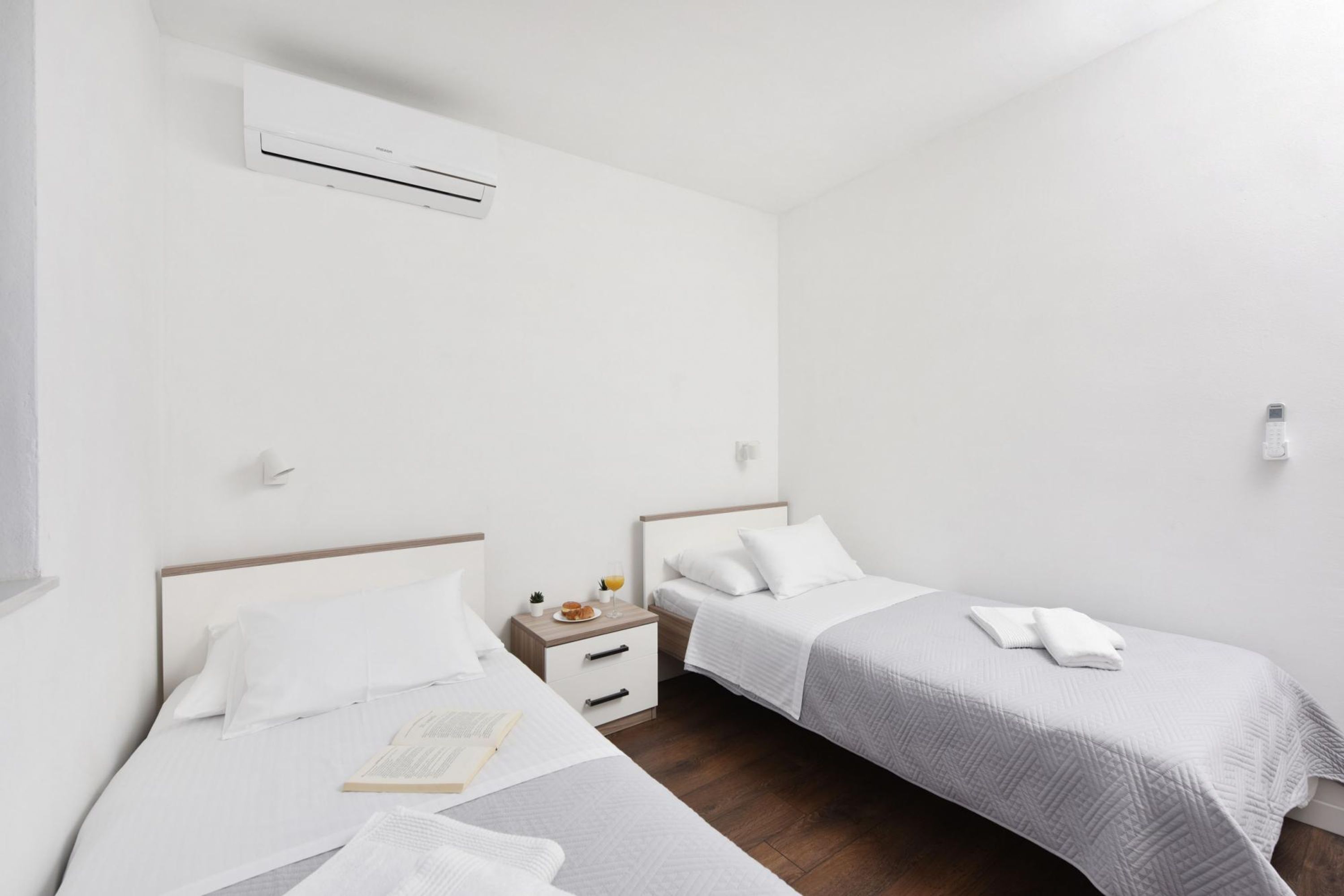 A hotel room with two single beds, a bedside table with a lamp, an air conditioning unit, and a small nightstand with a book and a glass of water.