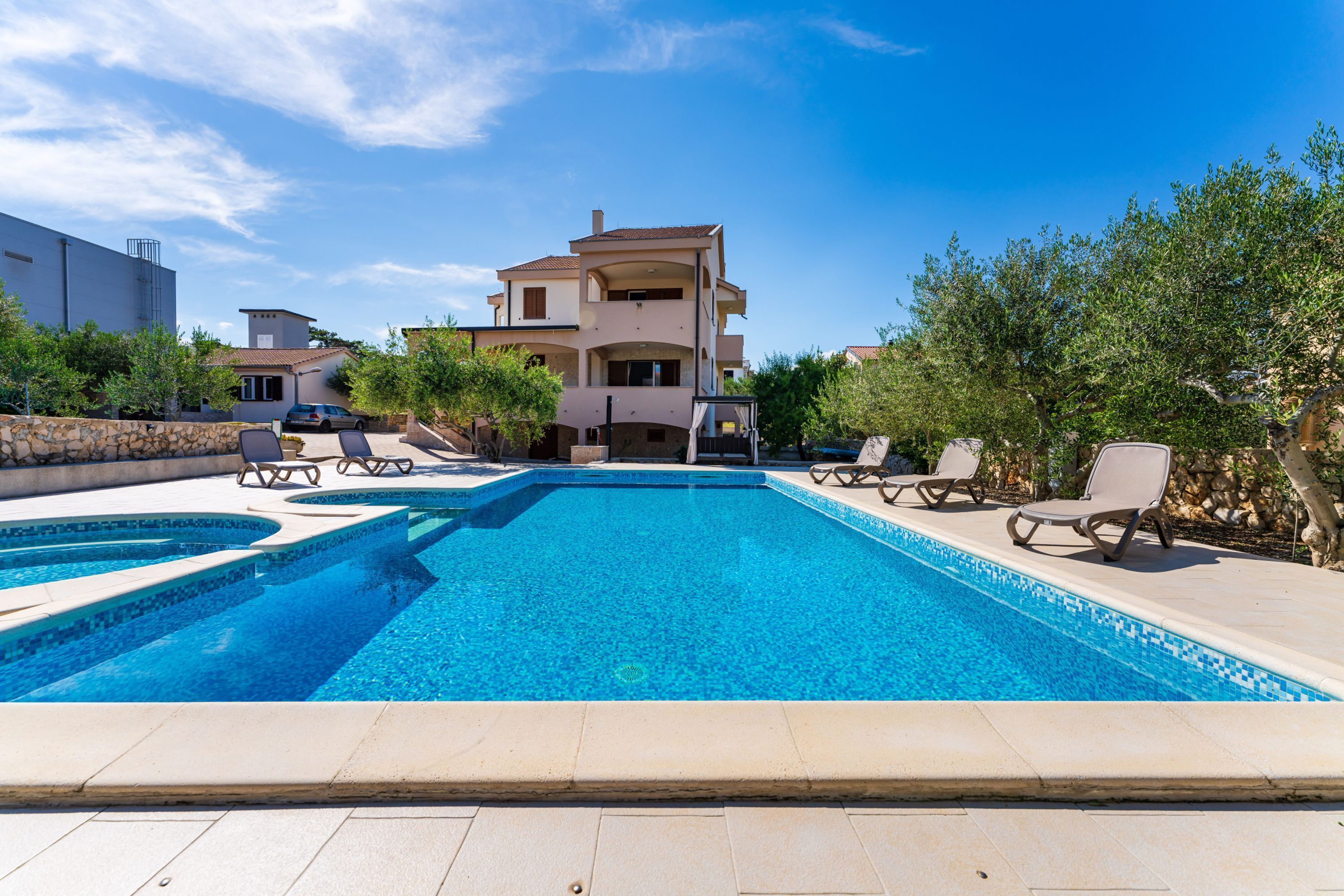 A swimming pool with lounge chairs, a stone wall, and a multi-story building in the background.