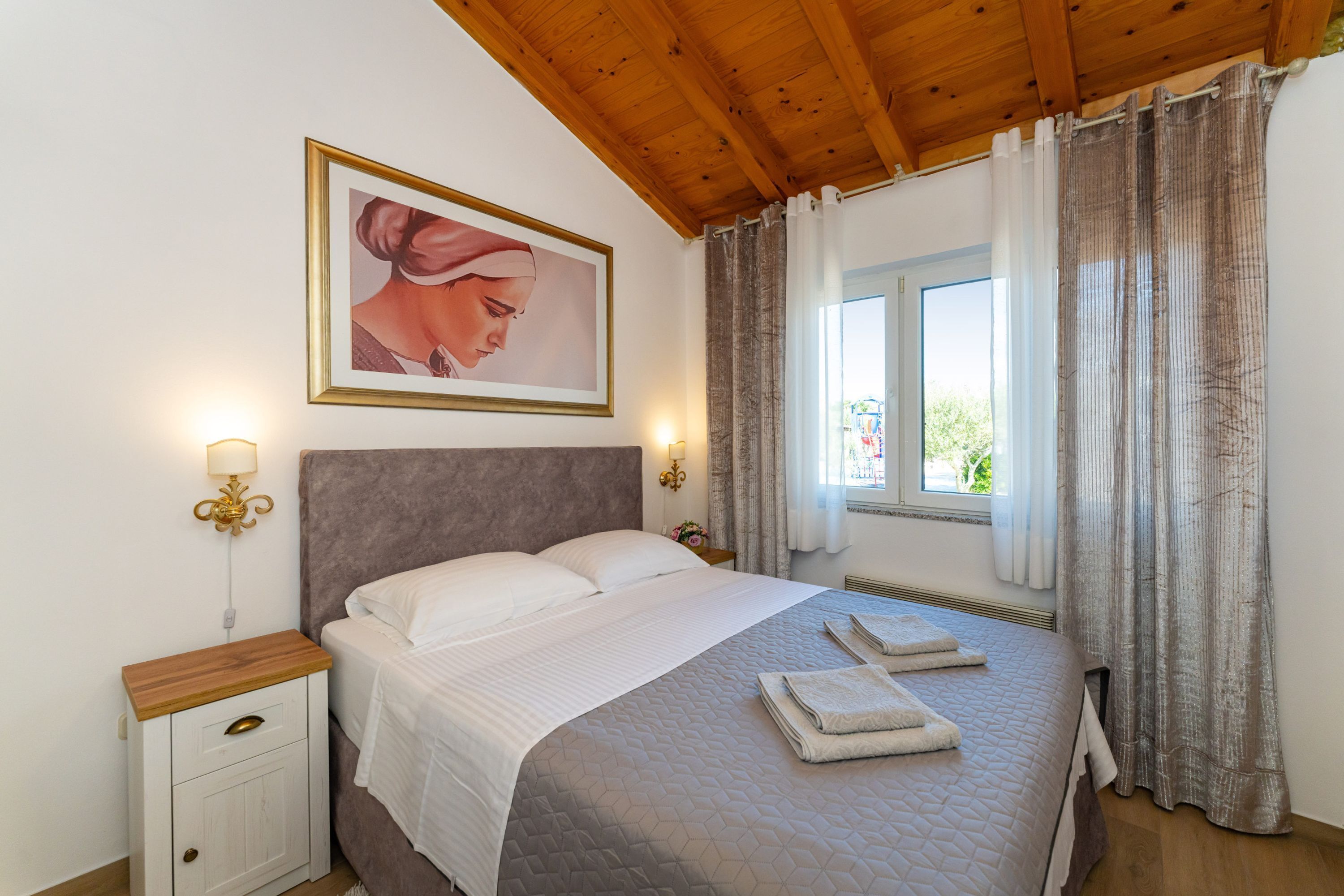 Un letto rifatto con copriletto trapuntato, un comodino in legno con cassetto, un quadro incorniciato appeso al muro e una finestra con tende.