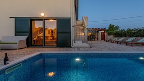 Une piscine avec une entrée constituée d’une porte vitrée, un espace extérieur aménagé pour s’asseoir et un escalier menant à une zone abritée.