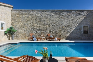 A pool area with stone walls, wooden lounge chairs, and a table with a vase of flowers and a bottle. - Villa Granar Zadarvillas (Benkovac)