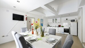 Ein moderner Essbereich mit einem weißen Tisch, gedeckt für vier Personen. Im Hintergrund ist eine Küche erkennbar, und an der Wand ist ein Flachbildfernseher angebracht.