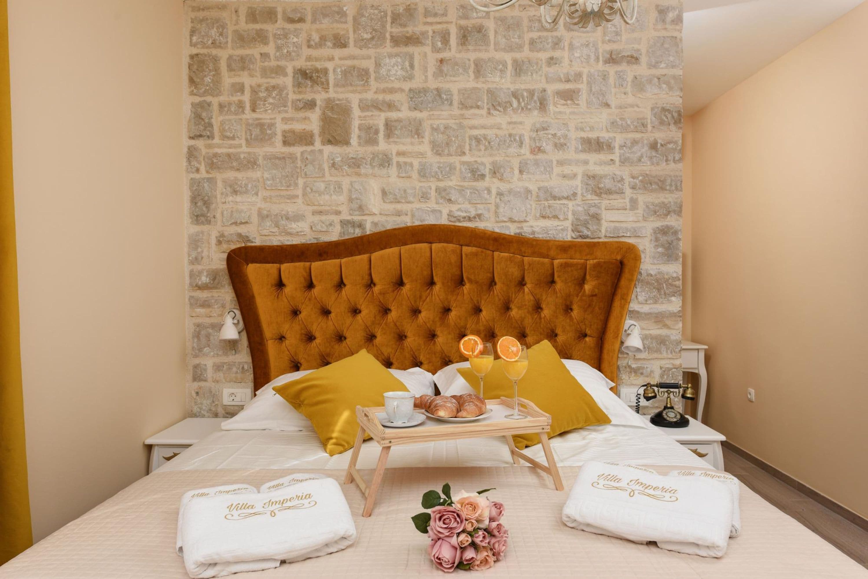 Un lit bien fait, agrémenté d’une tête de lit capitonnée, une tablette avec des croissants et du jus d’orange, et un bouquet de roses roses.
