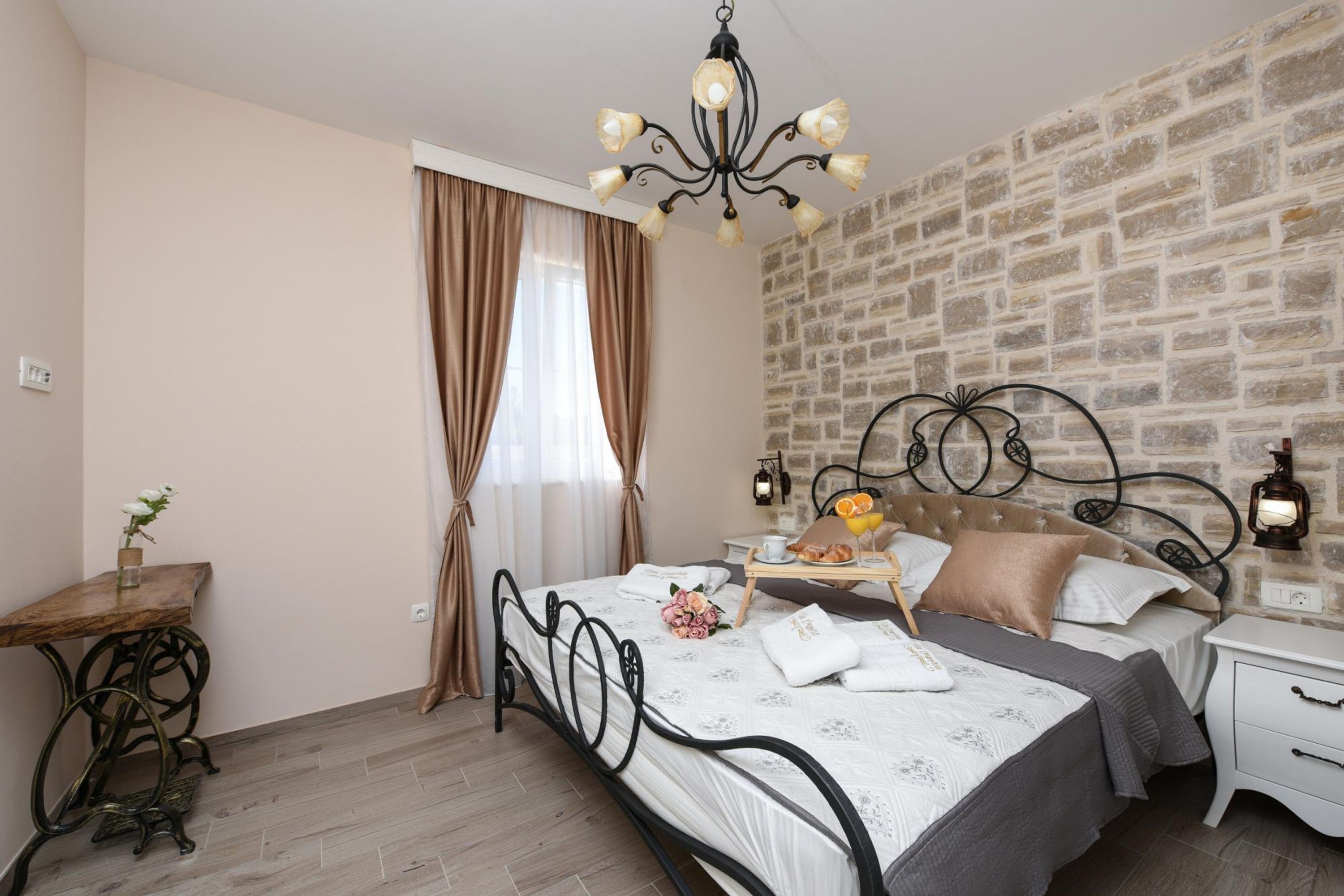 A bedroom with a brick wall, a bed with a metal frame, a bedside table, a nightstand, a chandelier, and a window with curtains.