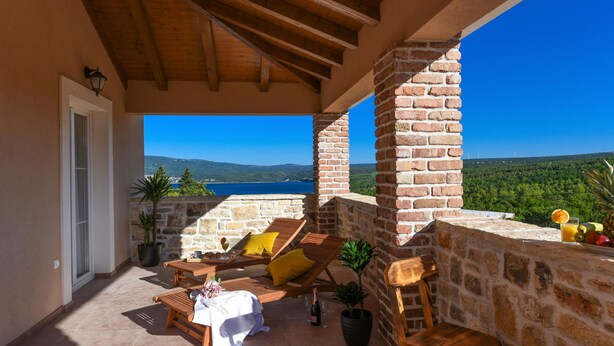 Une terrasse dotée de mobilier en bois, une table dressée pour le repas et une vue sur la mer et les collines.