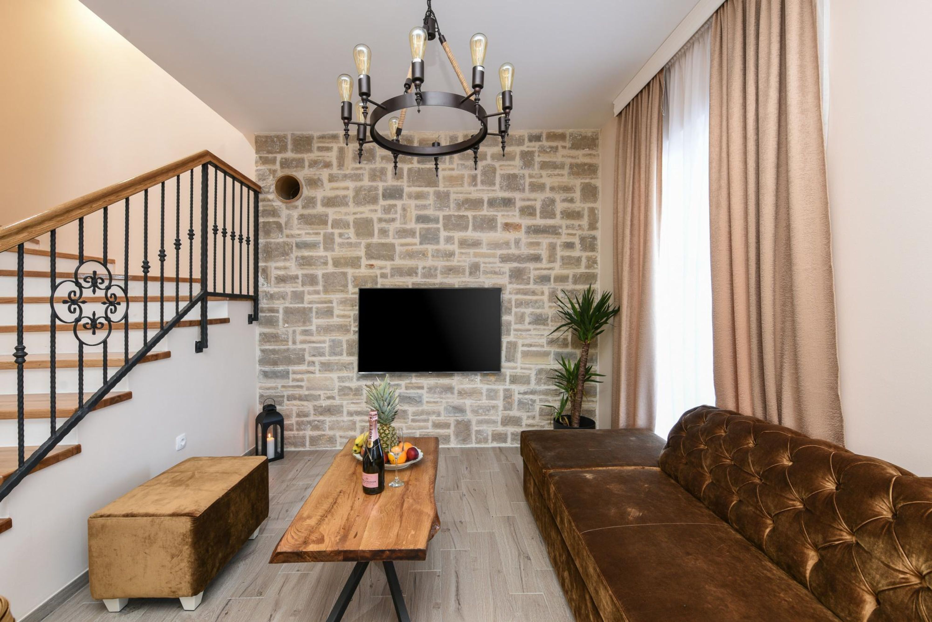 A living room with a stone wall, a flat-screen TV, a wooden coffee table with fruit and a bottle, a brown sofa, and a staircase with black railings.
