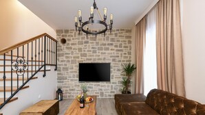 Un salon avec un mur en pierre, un téléviseur à écran plat, une table basse en bois sur laquelle se trouvent des fruits et une bouteille, un canapé marron et un escalier avec des rampes noires.