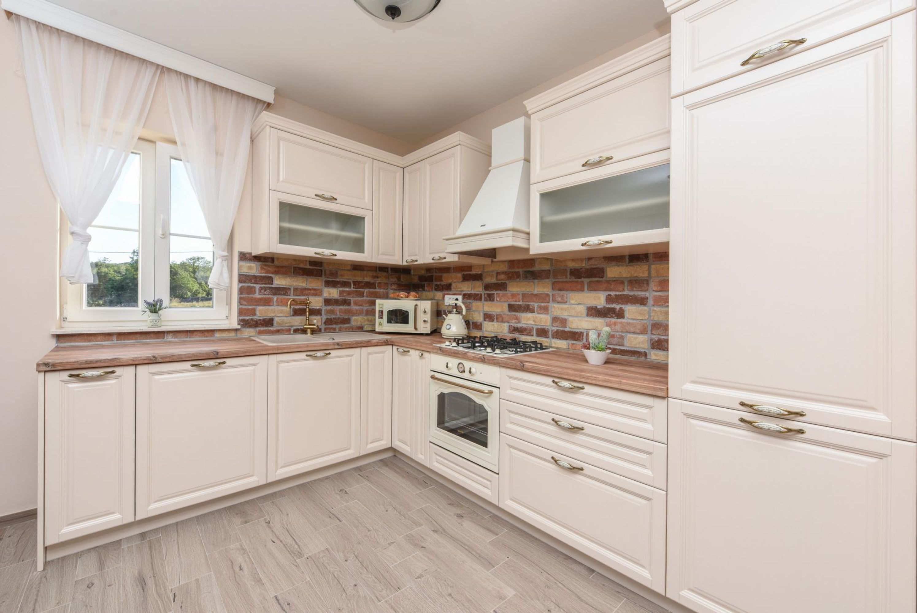 Una cocina con muebles blancos, salpicadero de ladrillo, microondas, estufa y ventana con cortinas ligeras.