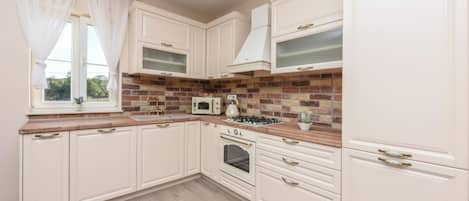Una cocina con muebles blancos, salpicadero de ladrillo, microondas, estufa y ventana con cortinas ligeras.