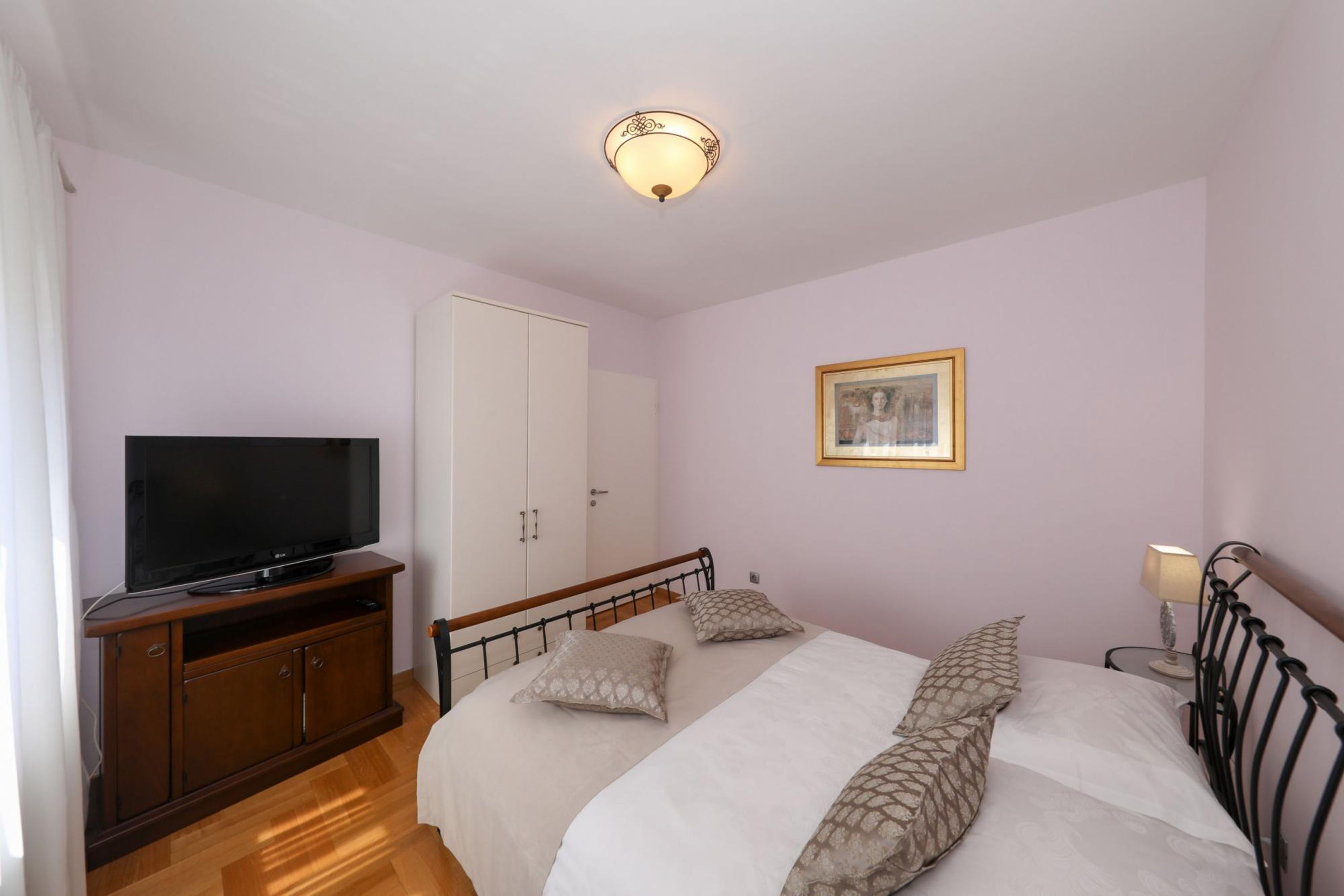 A bedroom with a bed, a TV on a wooden cabinet, a wardrobe, and a painting on the wall.