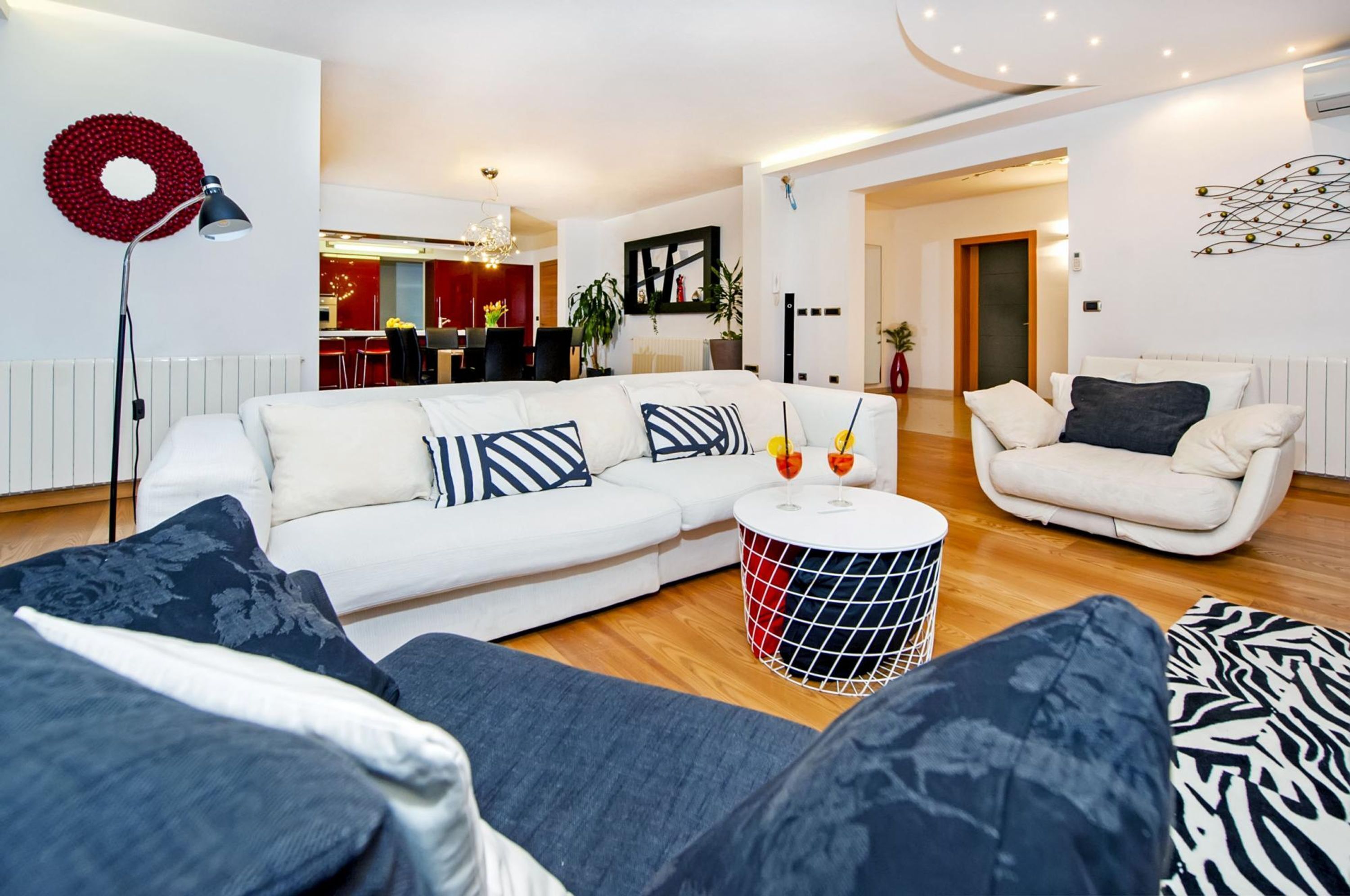 Ein modernes Wohnzimmer mit einem weißen Sofa, einem runden Couchtisch und einem weißen Sessel. Im Hintergrund befindet sich ein Essbereich mit roter und schwarzer Einrichtung.