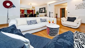 Un salon moderne avec un canapé blanc, une table basse ronde et un fauteuil blanc. On aperçoit à l’arrière-plan un coin repas décoré de rouge et de noir.