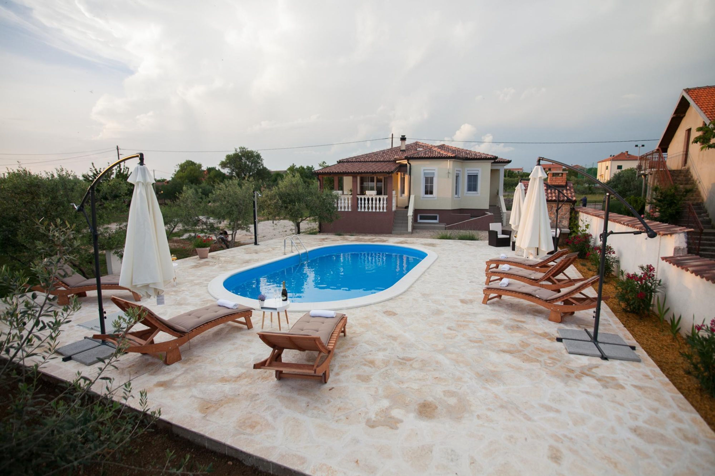 A swimming pool with lounge chairs and umbrellas, surrounded by a stone patio and a house with a tiled roof.
