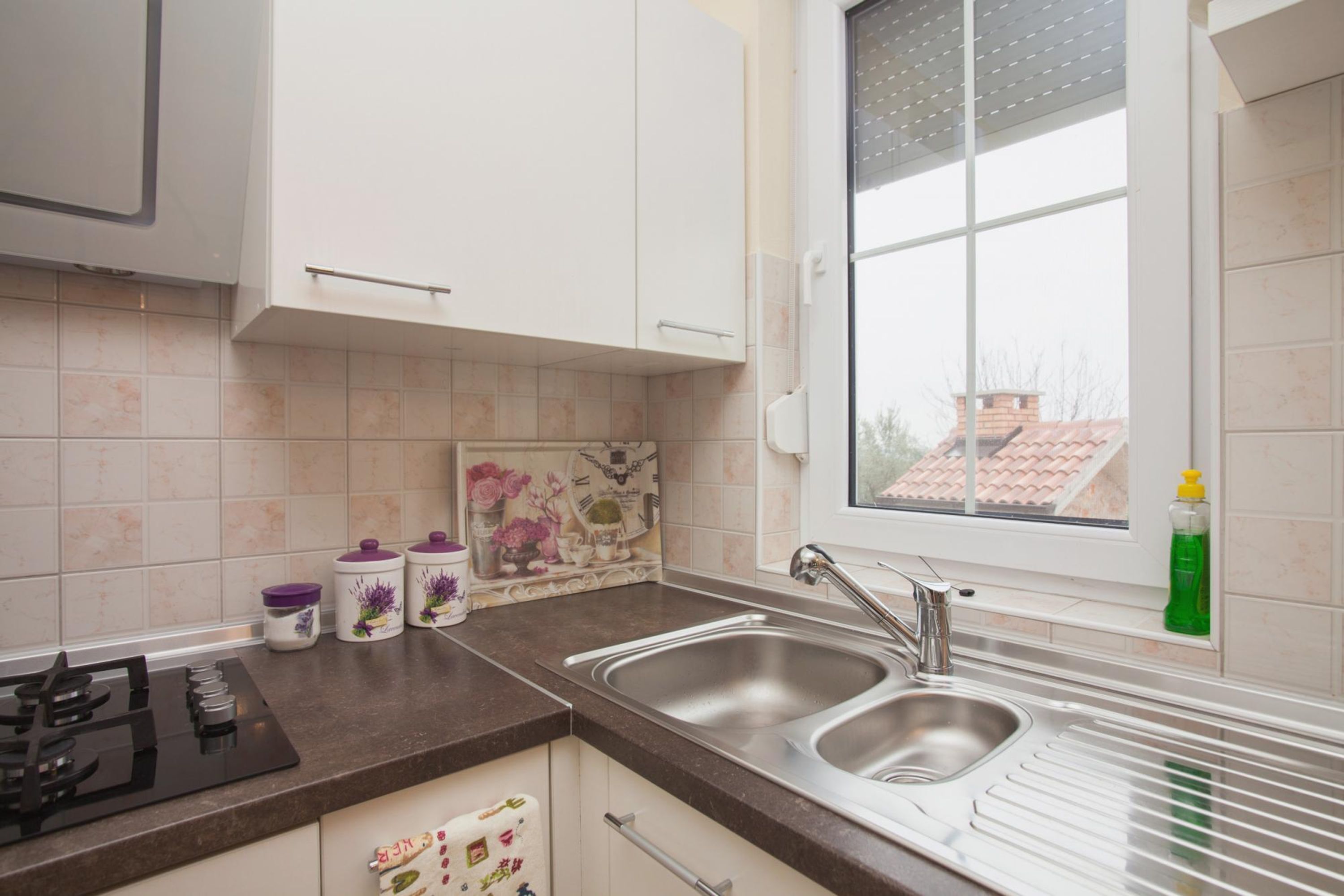 Une cuisine avec des meubles blancs, un évier en acier inoxydable et une fenêtre donnant sur une maison.
