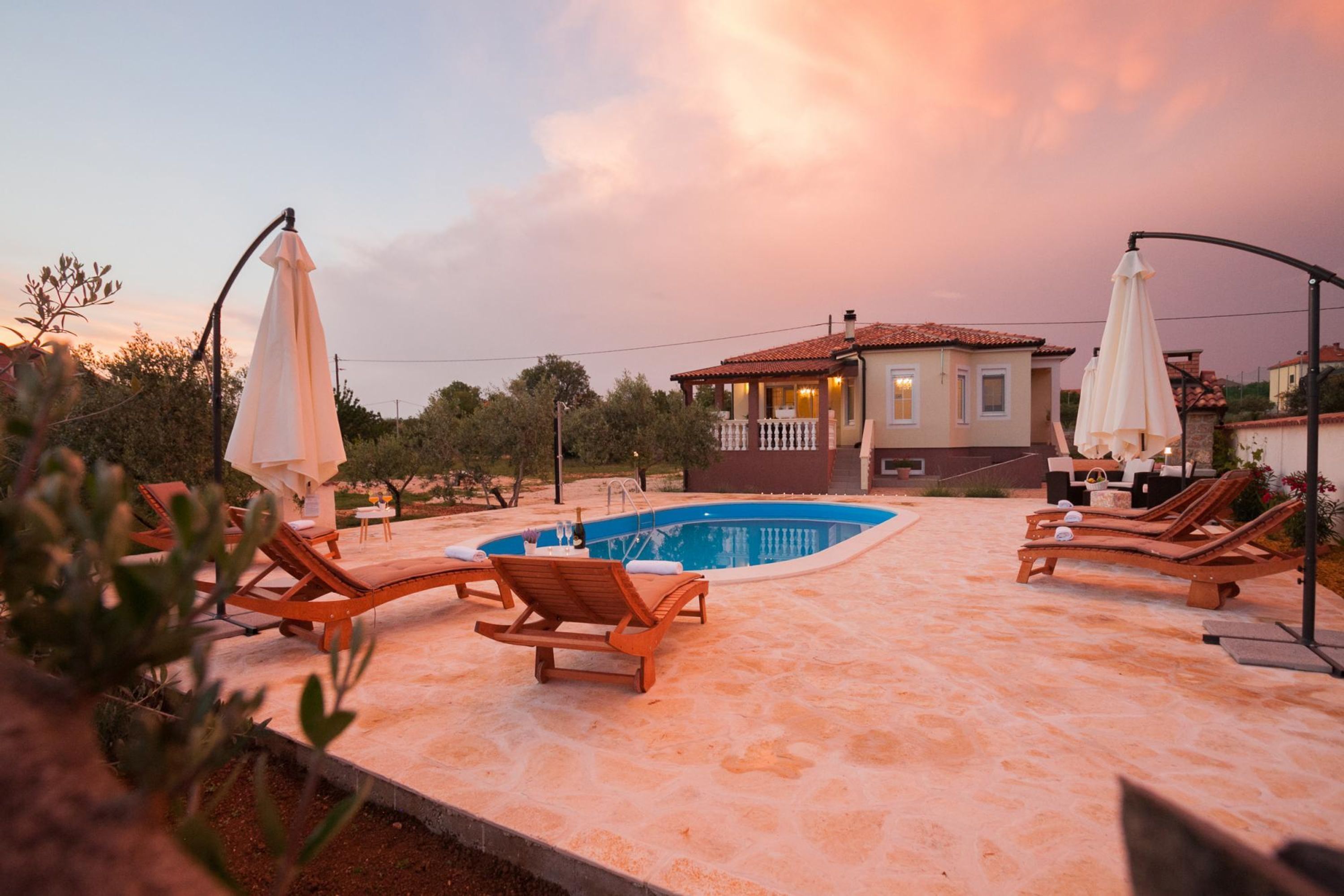 A pool area with lounge chairs and umbrellas, surrounded by a house with a tiled roof.