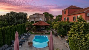 Un área de piscina con sillones de descanso y sombrillas, rodeada de vegetación y edificios.