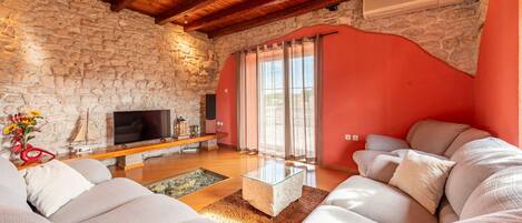 A living room with a stone wall, wooden ceiling, a flat-screen TV, a sofa, and a large window with curtains.