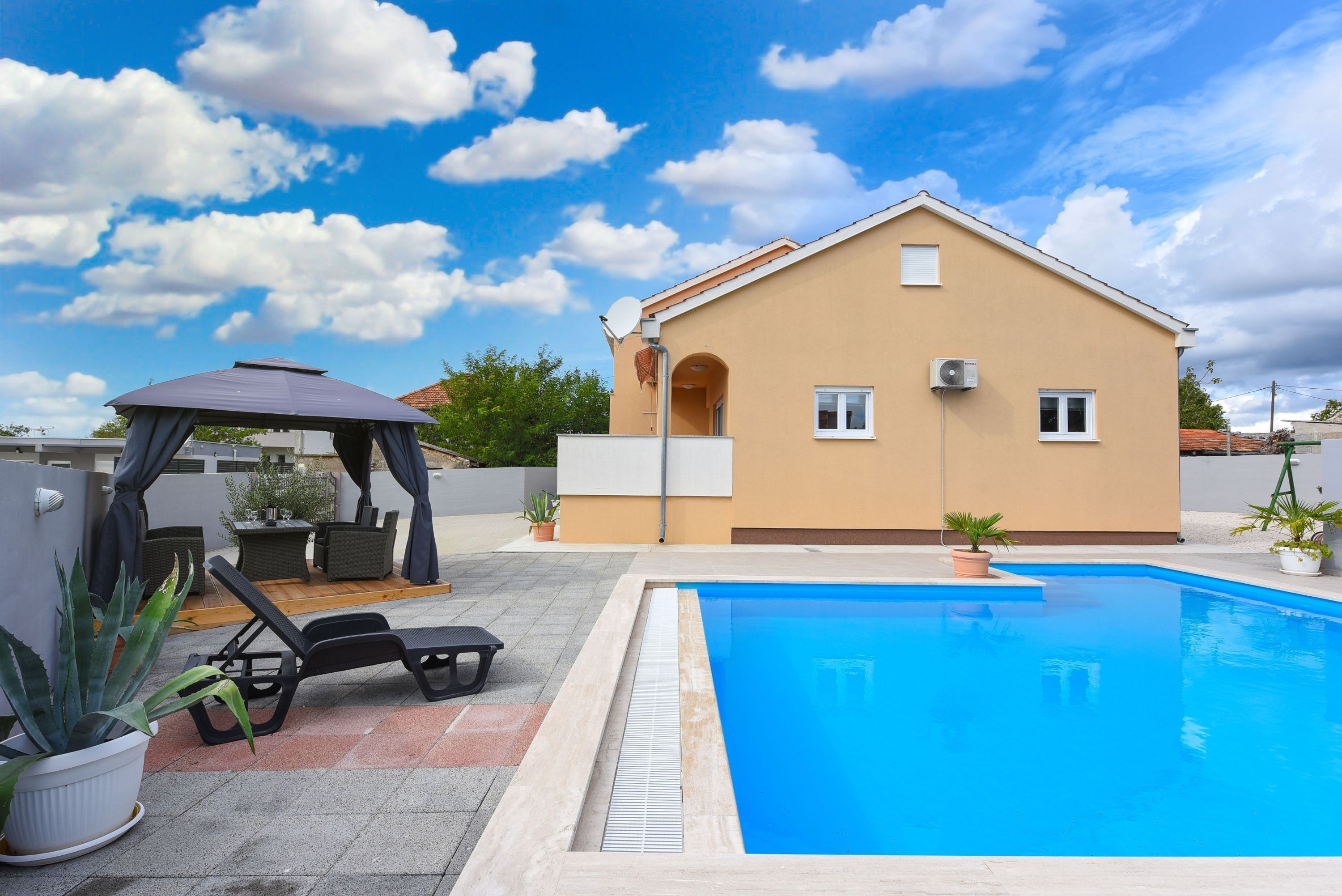 Une piscine avec des chaises longues et un kiosque, une maison beige avec une antenne parabolique et des plantes en pot.