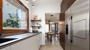A modern kitchen with a stainless steel refrigerator, wooden cabinets, a window with a view, and a dining area with a table and chairs. - Villa Antea Zadarvillas (Zadar)