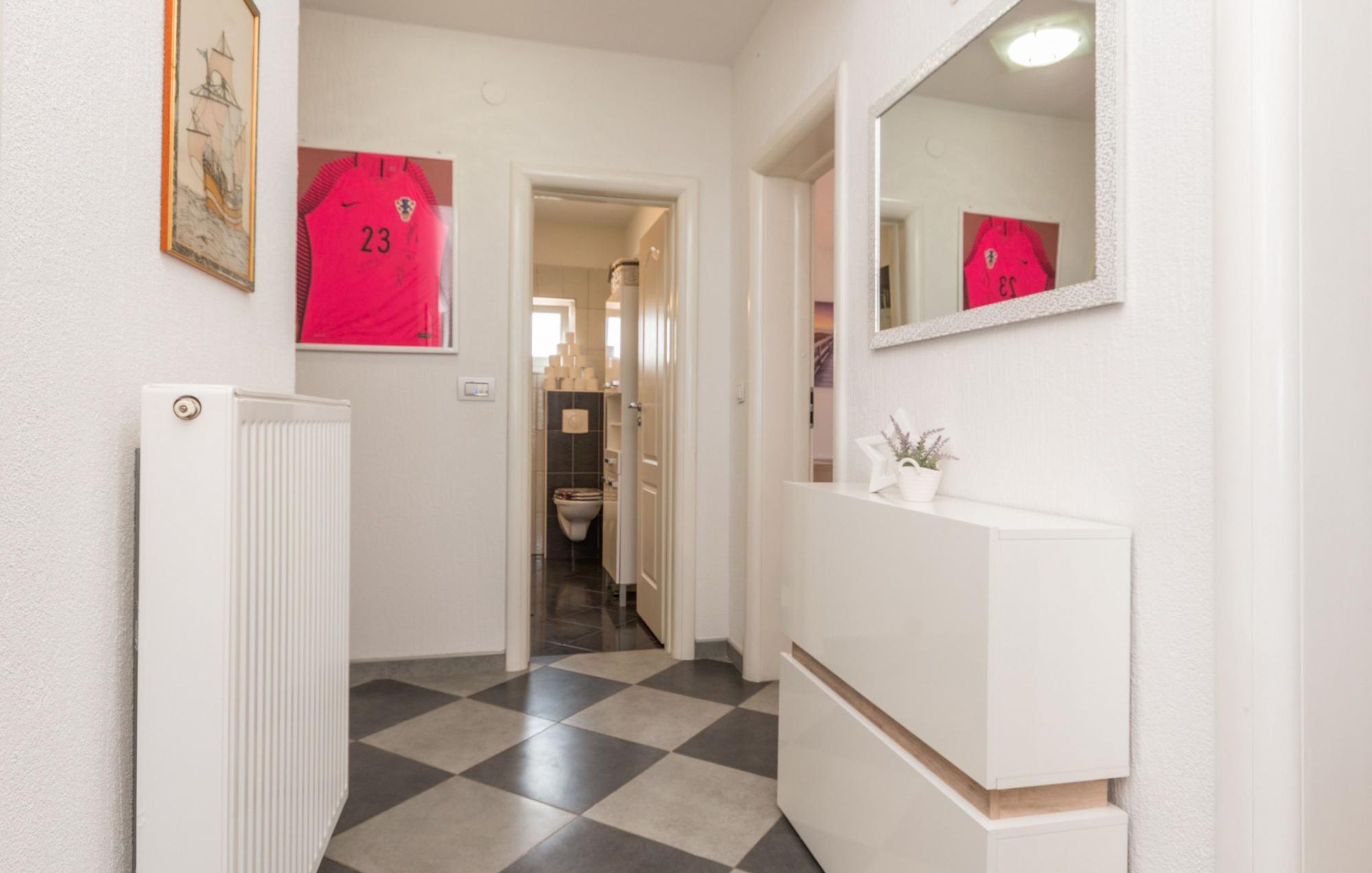 A hallway with a checkered floor, a radiator, a mirror, and a red sports jersey hanging on the wall.