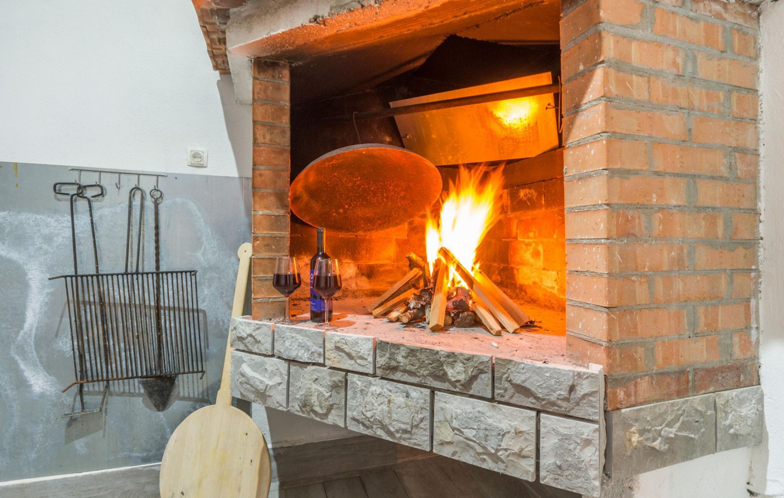 A brick oven with a fire burning inside, a hanging metal rack, and a wooden paddle.