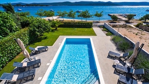 A rectangular swimming pool with a clear blue water, surrounded by lounge chairs and umbrellas, with a view of the sea and hills in the background. - Villa Roberta Zadarvillas (Sveti Filip i Jakov)