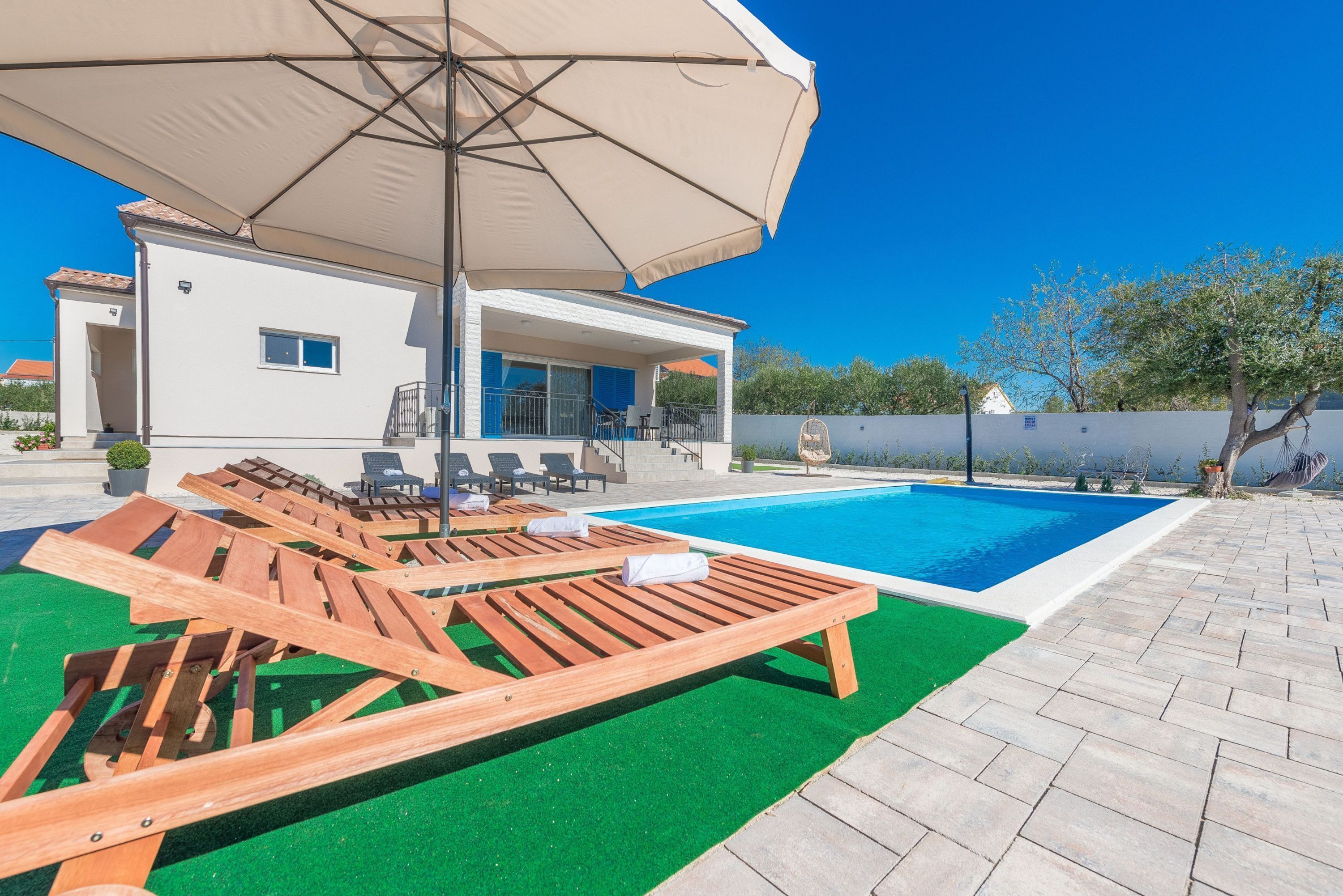 A pool area with wooden loungers and a large umbrella, a white building with a balcony, and a clear blue sky.