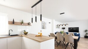 Una cocina moderna con armarios blancos, encimera de madera y un lavamanos integrado. Hay una zona de comedor con una mesa y sillas, y una sala de estar con sofá y televisor.